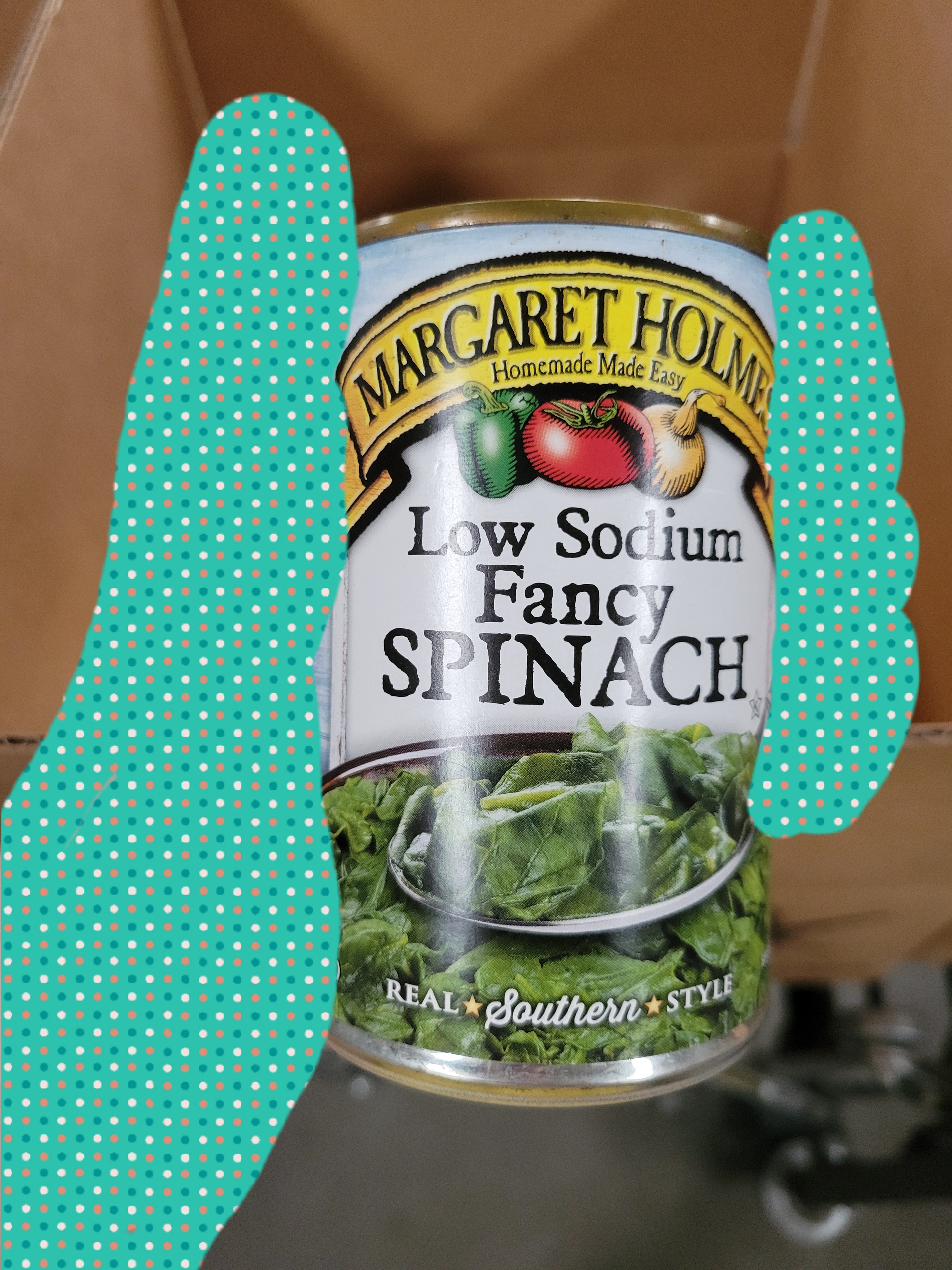 Hand holds can of fancy spinach in front of a cardboard box.
